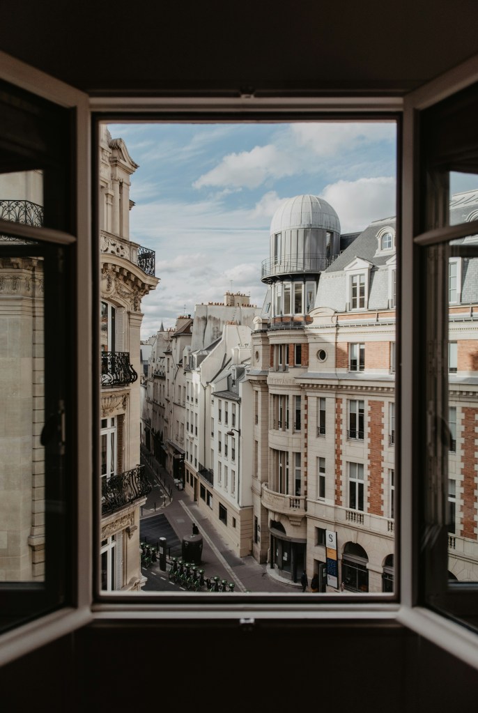 Vue sur une rue parisienne et ses immeubles depuis une fenêtre ouverte.